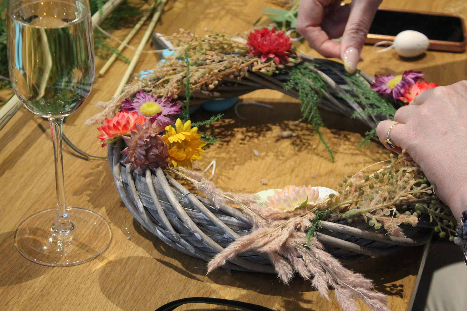 The photo shows a grey willow wreath with a glass of prosecco next to it. There are two hands working on the wreath base which is being adorned with pampas grass and colourful faux flowers.